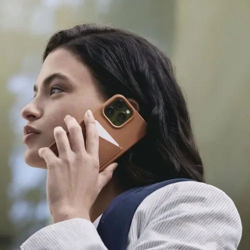 Helepruun nahkümbris kaardihoidjaga iPhone, mida hoitakse kõrva ääres; lähedalt on näha kolm kaameraobjektiivi ja valge kaardiserv.