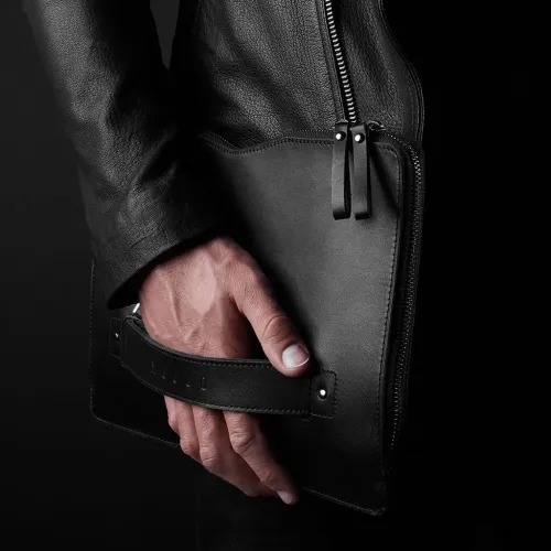 Black leather case with double pull tabs, visible edge zipper and back hand strap, held in hand against dark studio background.