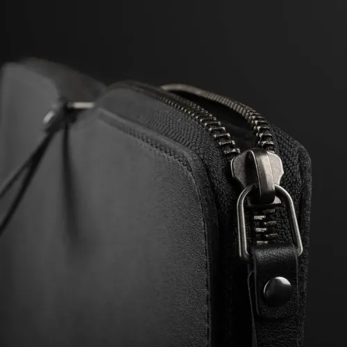 Mujjo MacBook futrālis no melnas ādas, tuvplāns augšējā stūrī ar raupju metāla rāvējslēdzēju, ādas ieliktni un redzamām šuvēm apkārt.